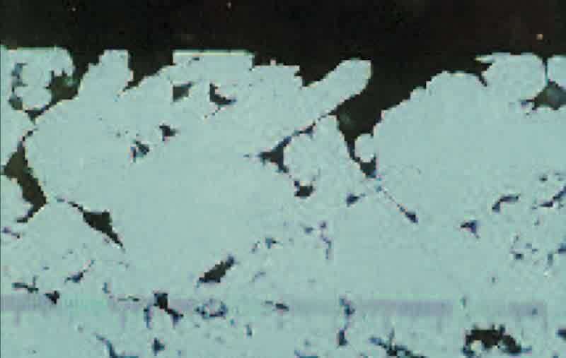 This is a cross-section of a plated part in the area evidencing the stain (400X). Note the nickel plates deep into the surface cavity and, because all sides of the nickel meet in the center, a microscopic “tunnel” is formed by the intersection. This tunnel leads to corrosion test failures, and cannot be eliminated by more plating. The absence of impregnation resin is evidenced by the nickel depositing deep into the surface pores.